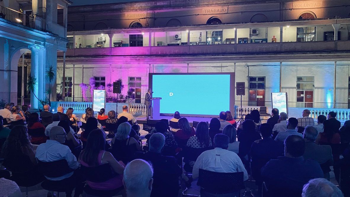 Público sentado frente al escenario y la pantalla en el patio del edificio, con iluminación azul y violeta.