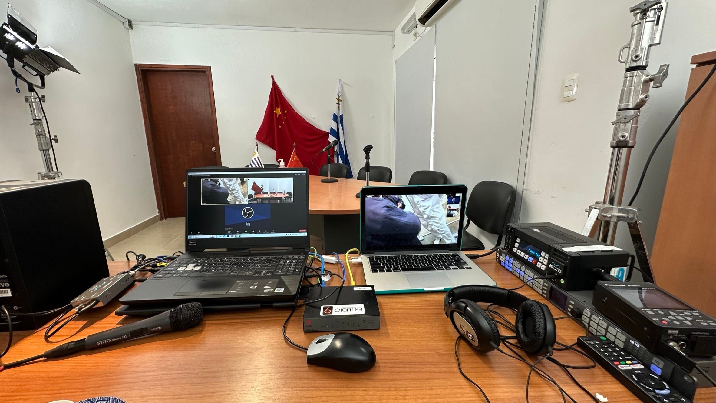 Notebooks, matriz de video, auriculares y micrófono armados sobre la mesa de la sala, con la videollamada en pantalla.