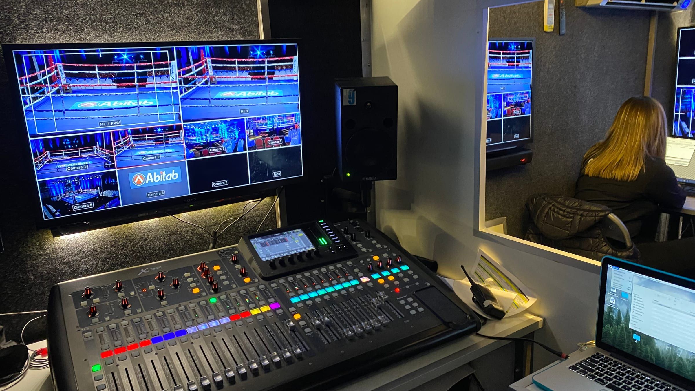 Consola de audio digital y monitor con la imagen del ring en el camión de transmisión, con una integrante del equipo detrás del vidrio