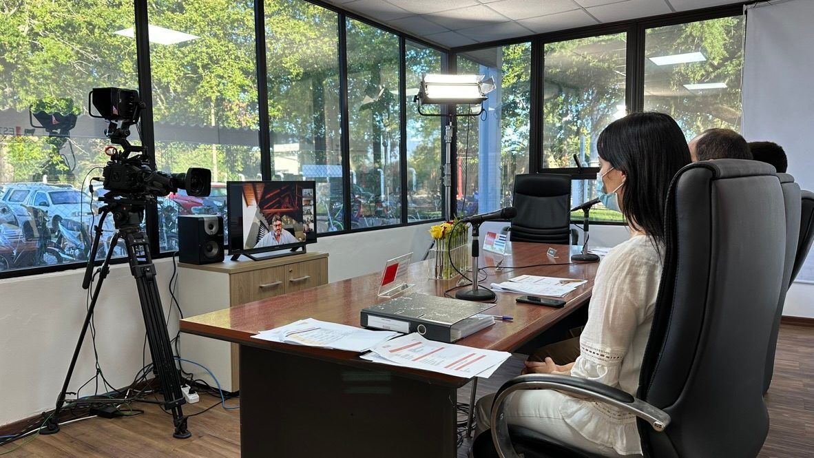 Cámara sobre trípode, luz y monitor de retorno frente a la mesa de la sala durante una auditoría virtual, con una participante de espaldas