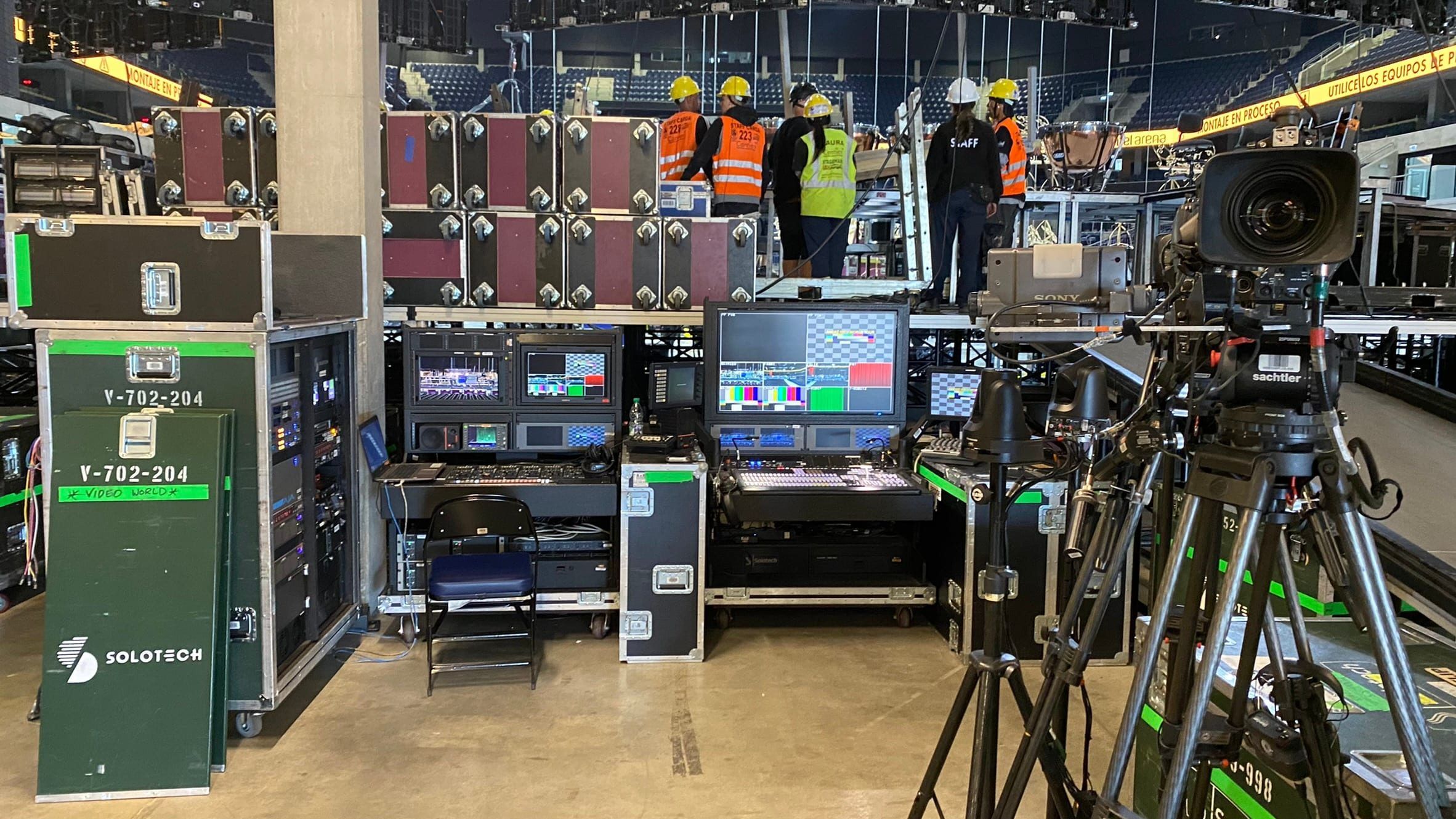 Cámara de estudio sobre trípode junto a las mesas de control y monitores, con el equipo técnico trabajando al fondo del Antel Arena