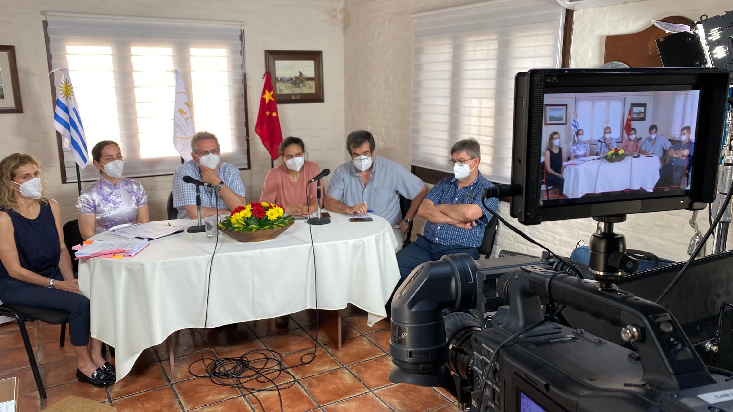 Mesa con delegados entre las banderas de Uruguay y China, con una cámara y su monitor de retorno en primer plano durante una auditoría virtual.